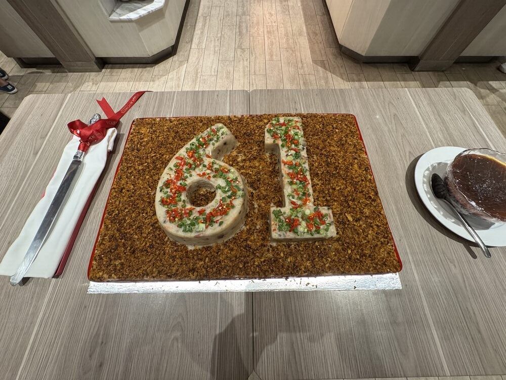 Steamed carrot cake decorated with the numerals 61 in white, on a wooden board with a cake knife beside it