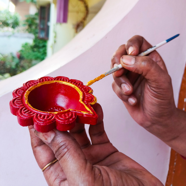 Hands painting a red clay diya lamp with a fine brush