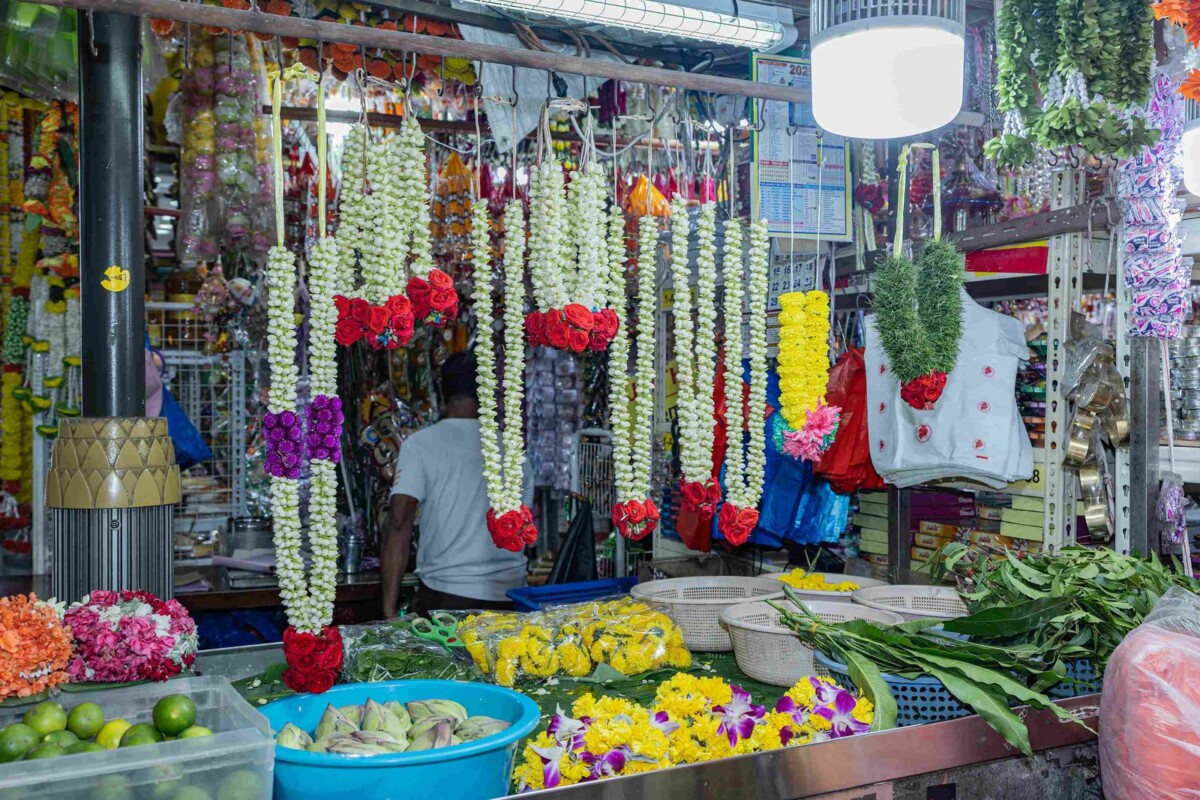 A flower stall interior with jasmine and red rose garlands hanging from a rod, a counter of loose marigolds, limes and lotus buds, and a worker facing away