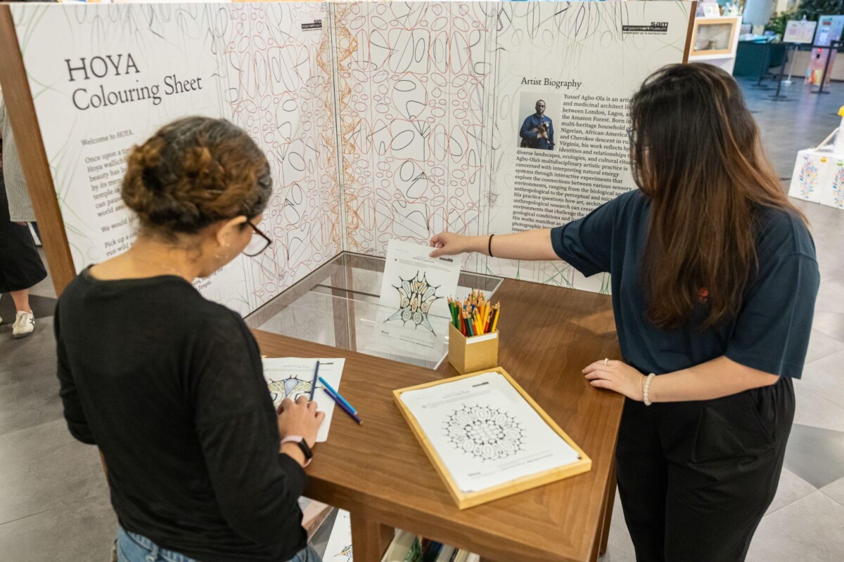 Two visitors colouring HOYA colouring sheets at a wooden station, with the artist biography panel behind them
