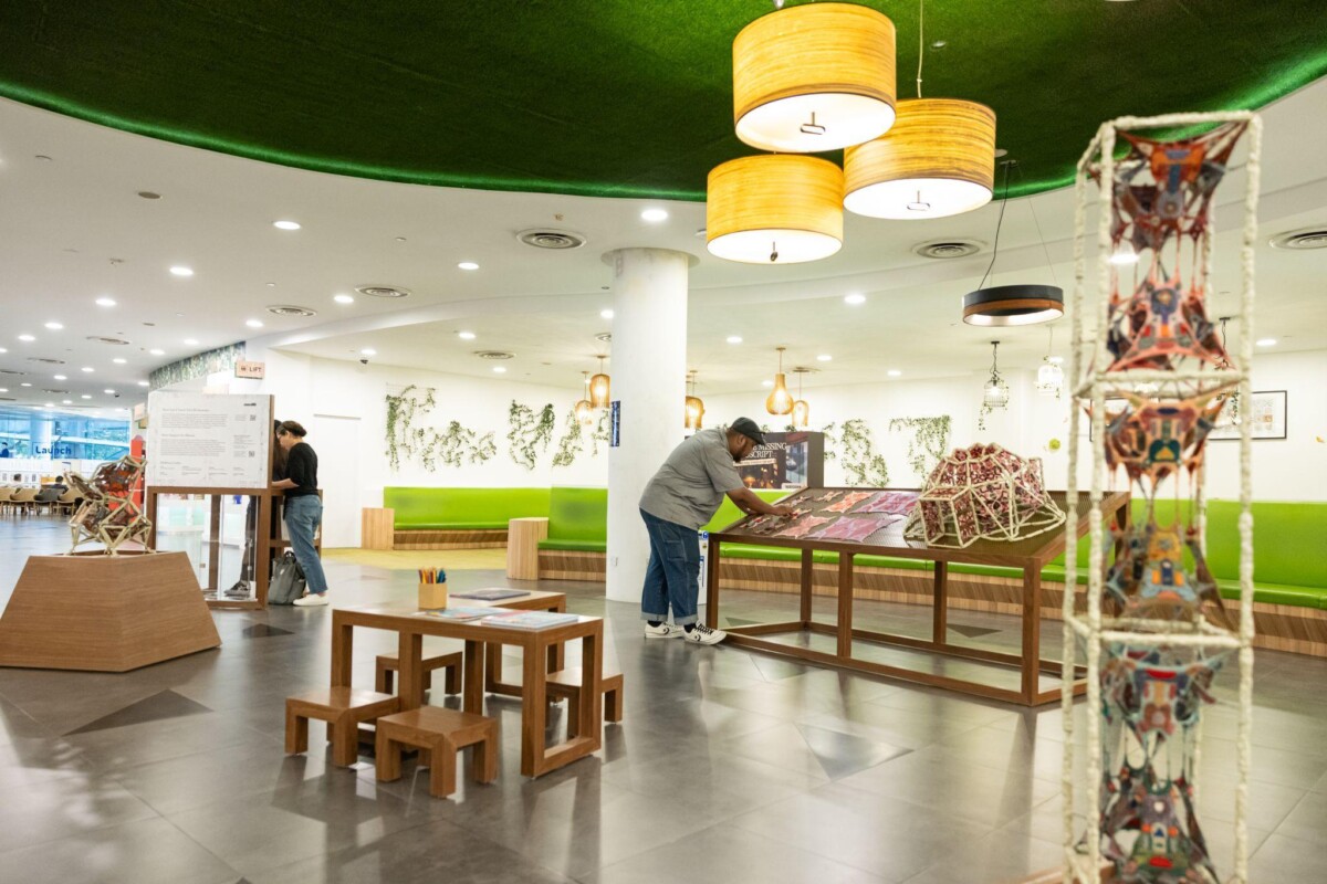 Exhibition view of Proposal for HOYA: 5 Lungs Temple at Jurong Library, with a visitor leaning over the display table and a tall knitted tower in the foreground