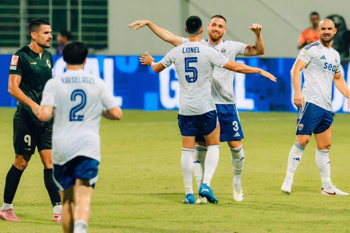 Lion City Sailors players in white celebrate on the pitch as a Tanjong Pagar player walks away