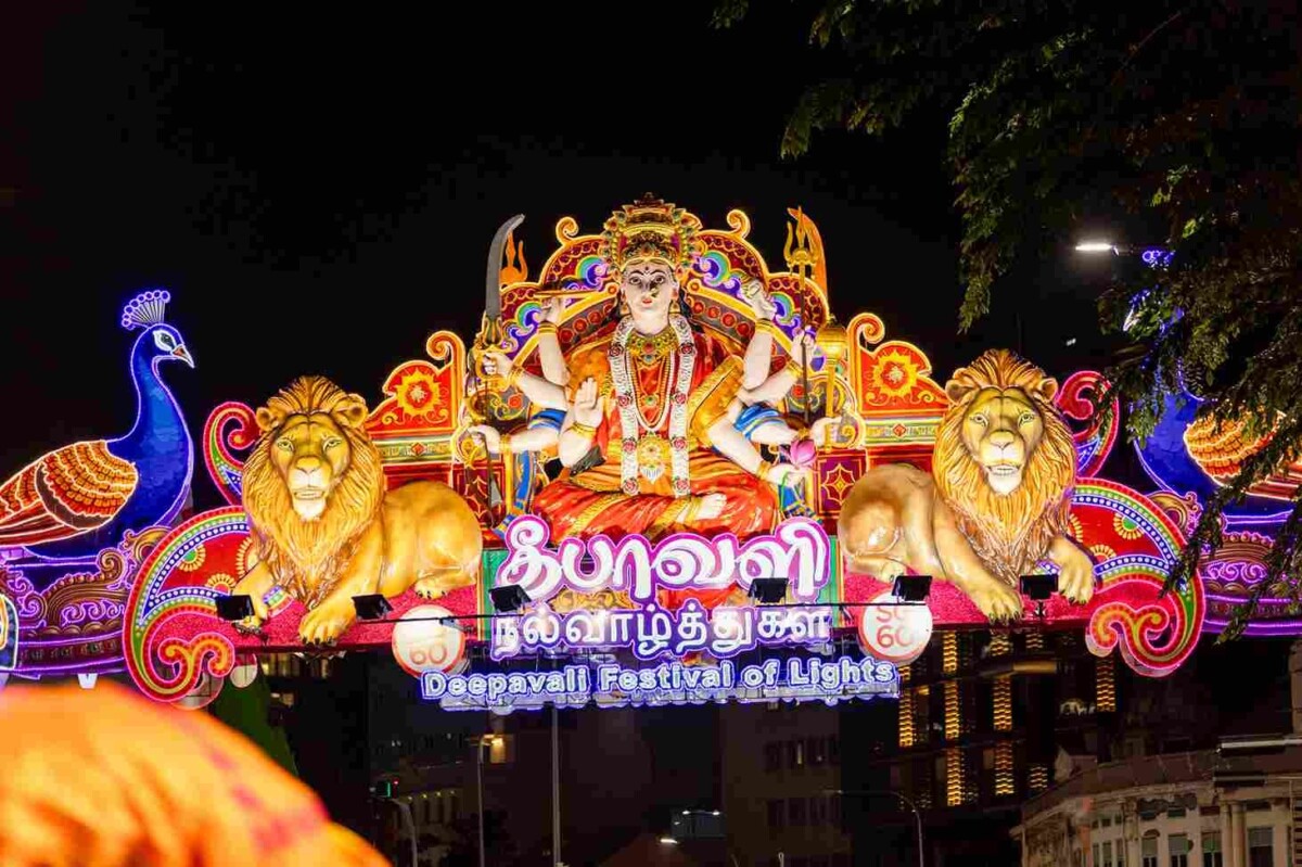 A lit Deepavali arch over a street in Little India at night, with a seated goddess figure flanked by two golden lions and blue peacocks at either end