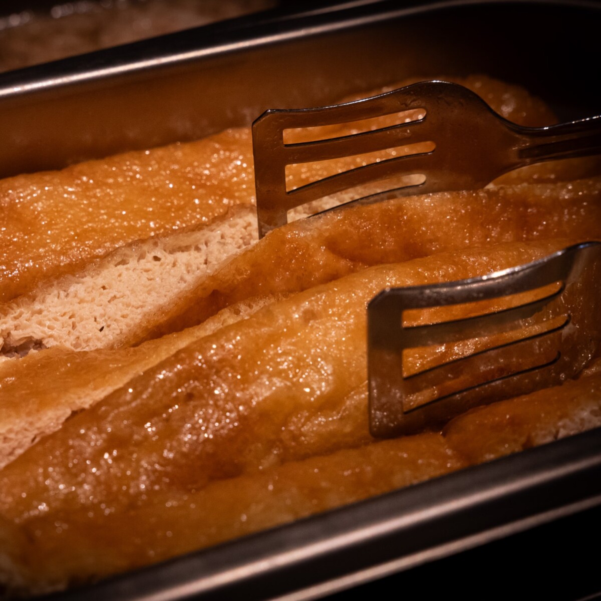 Tongs lifting large sheets of Japanese fried beancurd from a tray at Nekobushi