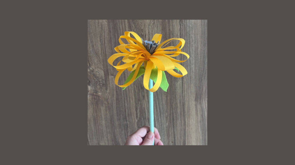 Hand holding a yellow folded paper flower on a green stem against wood