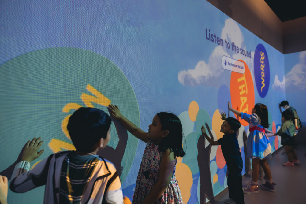 Children press their hands on a colourful interactive poetry wall reading Listen to the sound at Punggol Regional Library