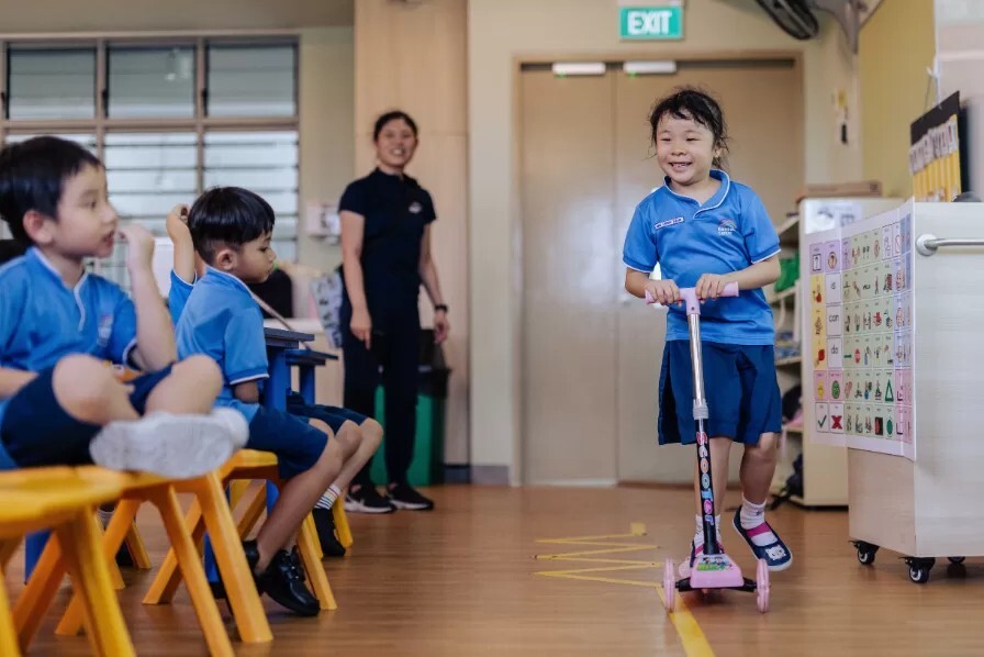 A pupil rides a scooter in a Rainbow Centre SPED classroom as classmates and a teacher watch