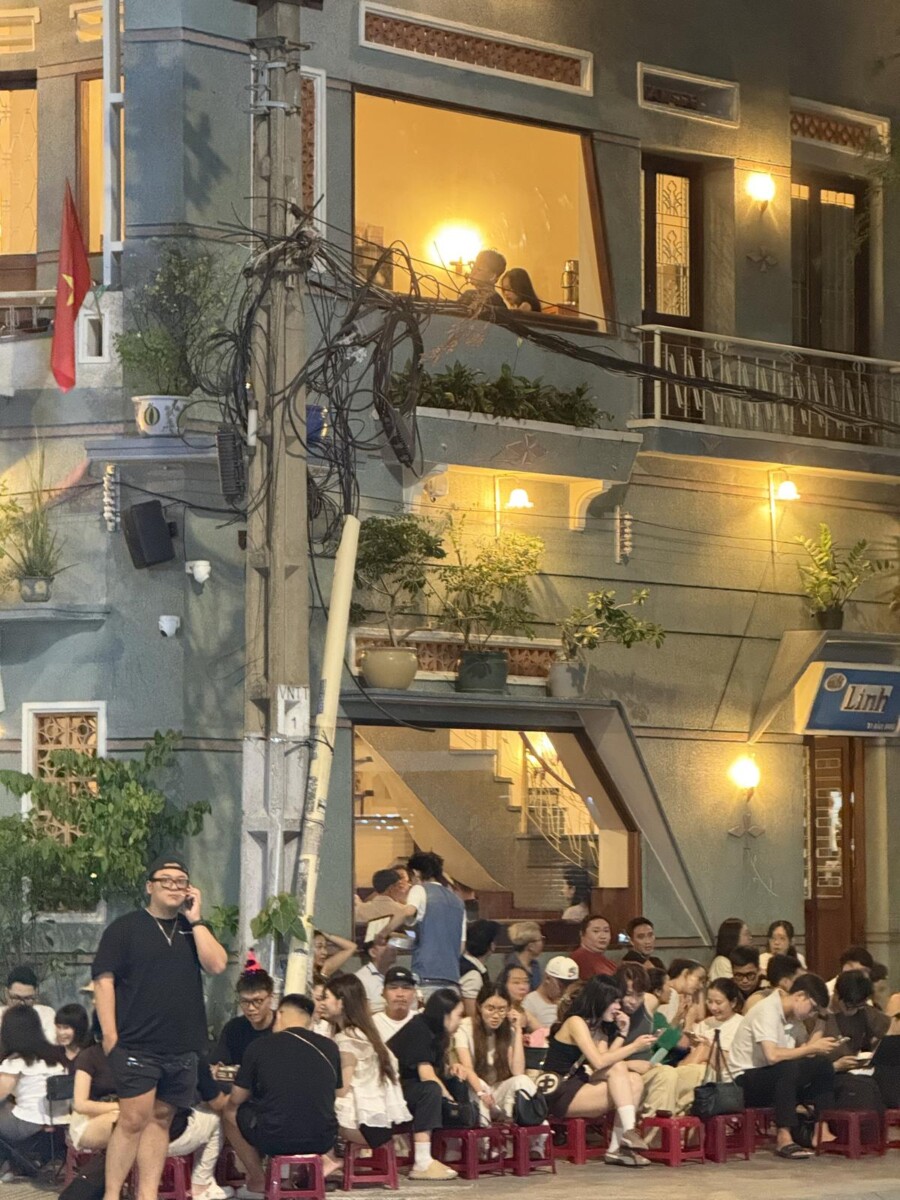 Saigon nightlife scene with youngsters packed on tiny plastic stools outside a colonial-style shophouse, a couple on the balcony above