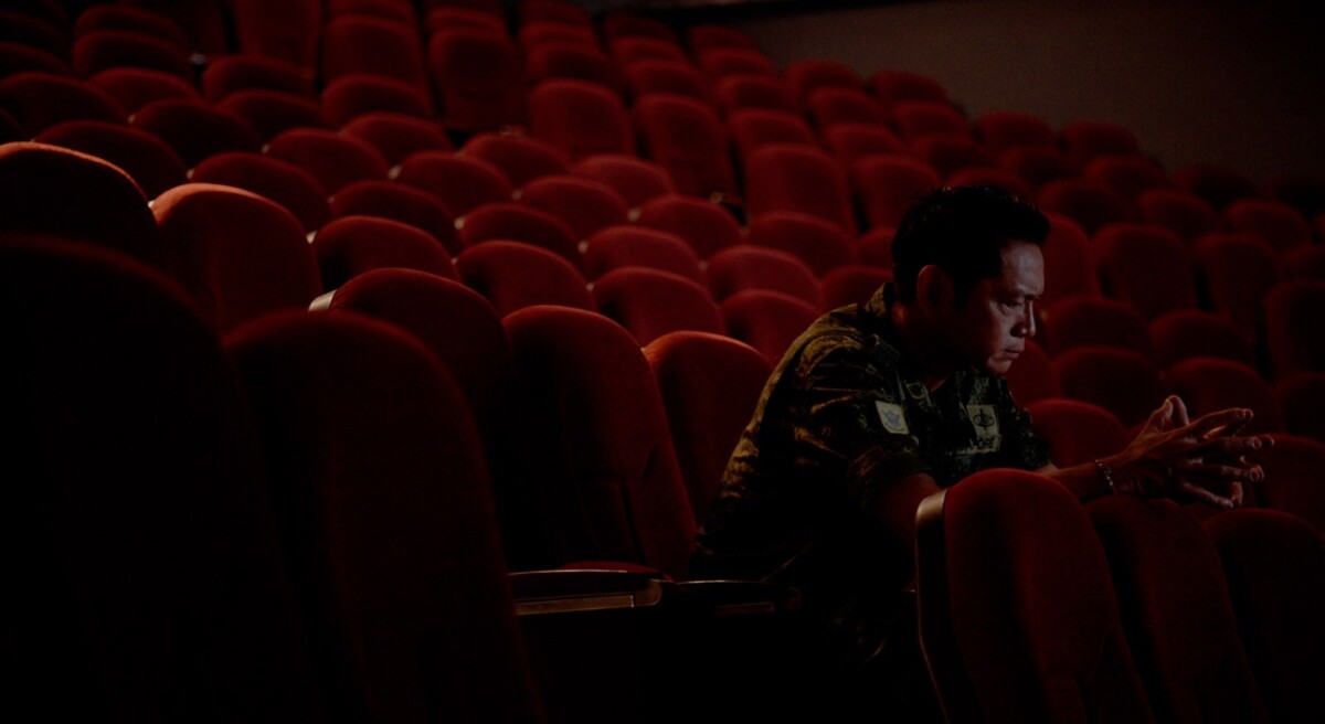 Army warrant officer sitting alone in rows of red seats in a still from Encik