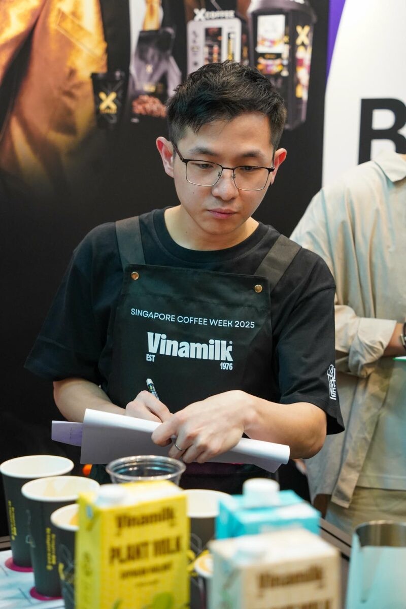 Barista in a Singapore Coffee Week 2025 apron writing notes at a booth with plant milk cartons and cups