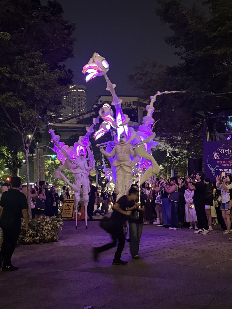 Wide shot of several illuminated pink and purple stilt-walking bird puppets from Birdmen towering over a street crowd at night, with a Singapore Night Festival banner in the background