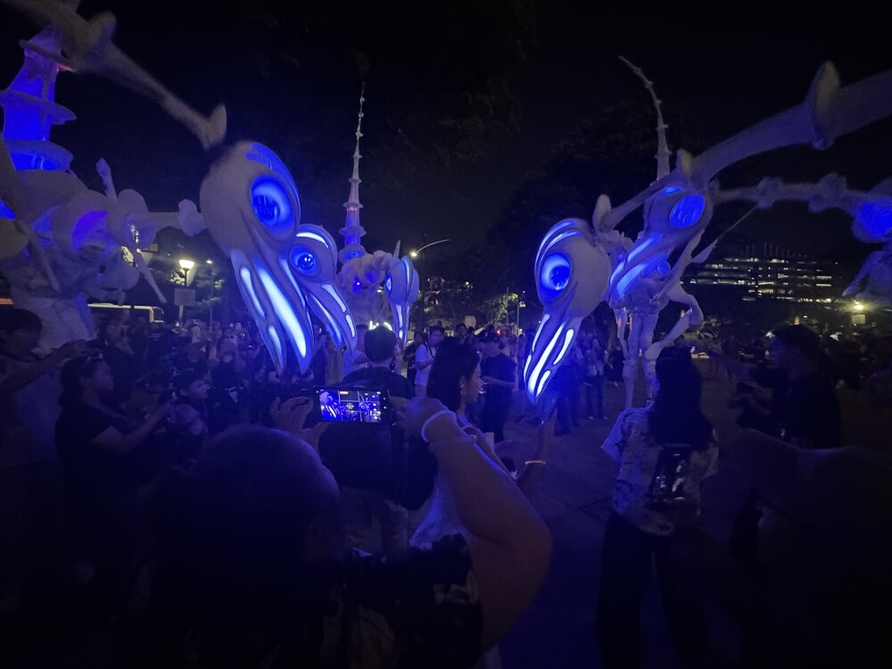 Close-up of a Birdmen stilt puppet's glowing blue skull-like head with illuminated eyes, seen from below at night