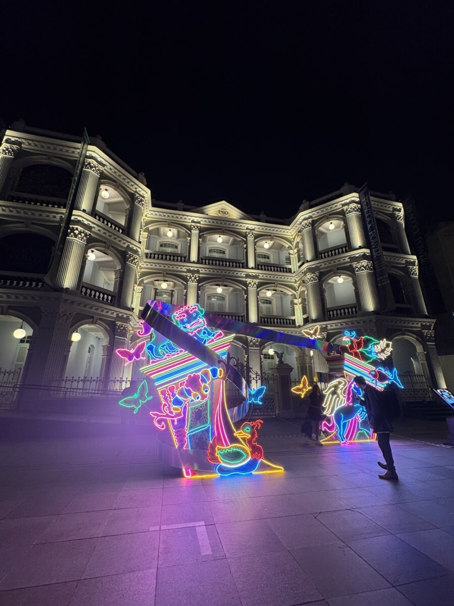 A colourful neon-outlined sculpture depicting a peacock and wayang-style warrior figures, mounted in front of a heritage building with arched colonnades