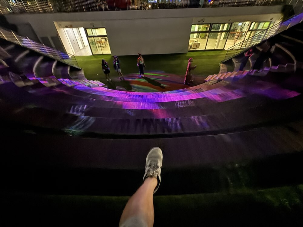 View from the top of a wooden amphitheatre lit in purple and pink, looking down at performers standing on the stage below, with trees and buildings framing the space