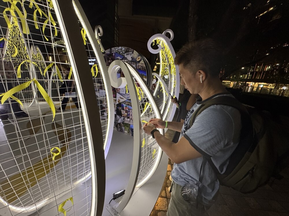 A visitor tying a yellow ribbon onto a large illuminated white wire-mesh petal-shaped sculpture at night