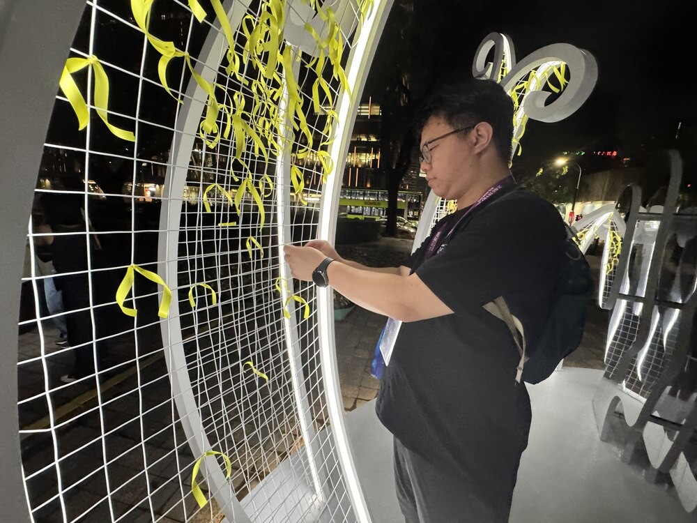 Another visitor tying a yellow ribbon onto the wire-mesh petal sculpture, with rows of previously tied yellow ribbons visible behind him