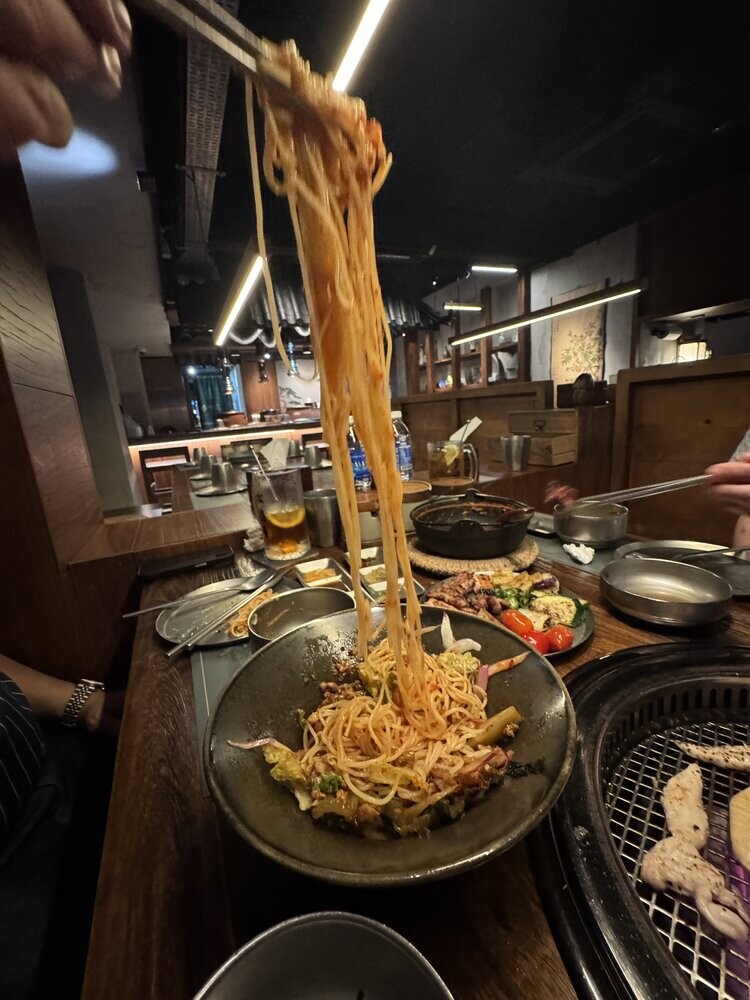 A long stretch of gochujang-tossed bibim noodles being lifted with chopsticks above the serving bowl, table spread visible in the background