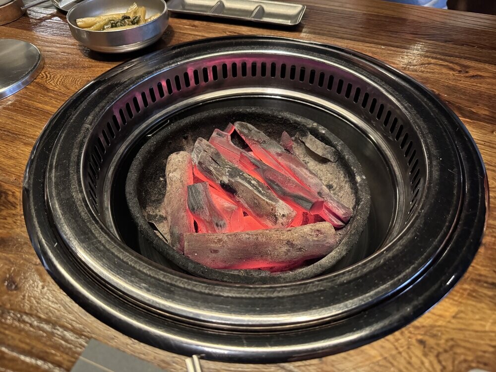 Close-up of glowing charcoal briquettes inside a tabletop grill, with a small side dish of banchan visible on the table