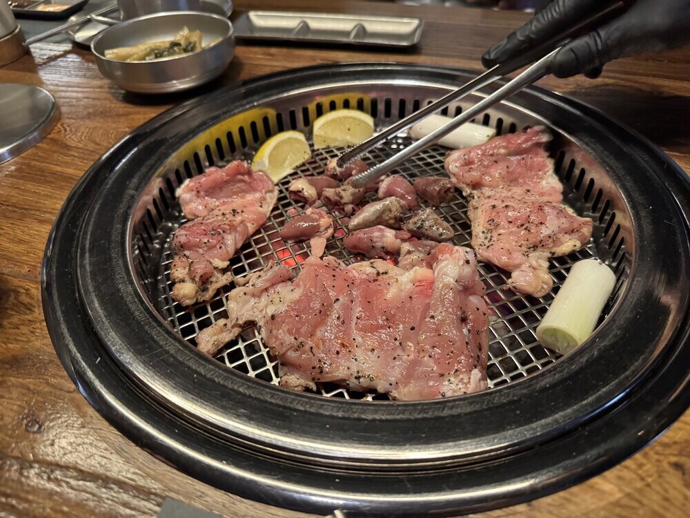 Chicken hearts and gizzards being turned with tongs on a charcoal grill, alongside marinated thigh and neck fillet cuts