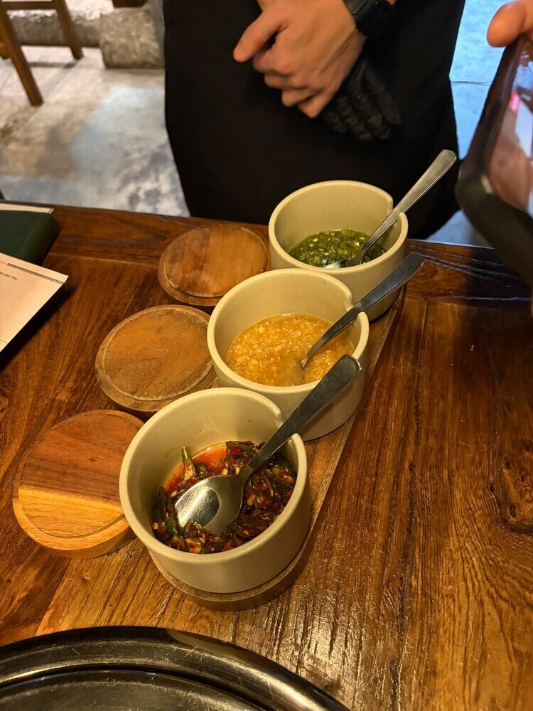 Three small ceramic pots of dipping sauces, green herb, garlic and red chilli, each with its own spoon, set on a wooden board