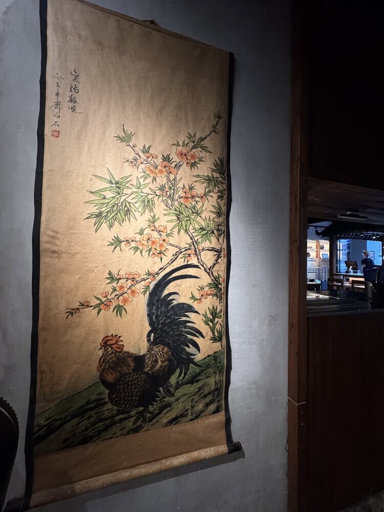 Traditional painted scroll of a rooster among peach blossoms and bamboo, hanging on a concrete wall near the restaurant entrance