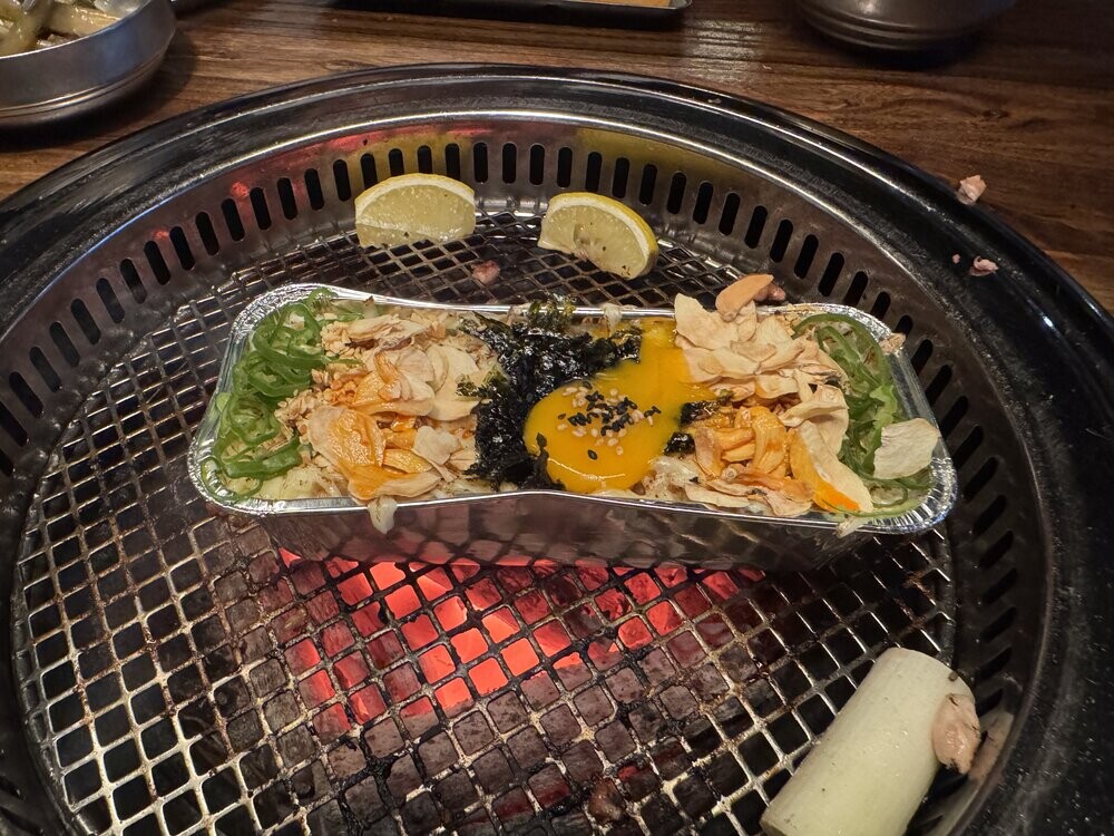 Uiseong Garlic Fried Rice cooking in a foil tray on the grill, topped with a raw egg yolk, shredded nori, garlic chips and scallions