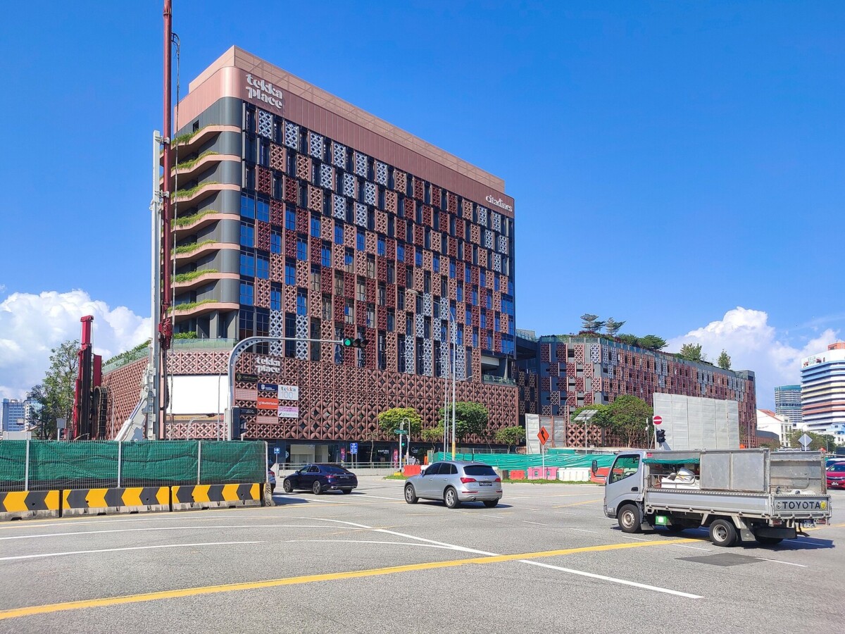 The exterior of Tekka Place, a modern tower with copper and grey geometric screens above a red-brown latticework podium, seen across a road junction