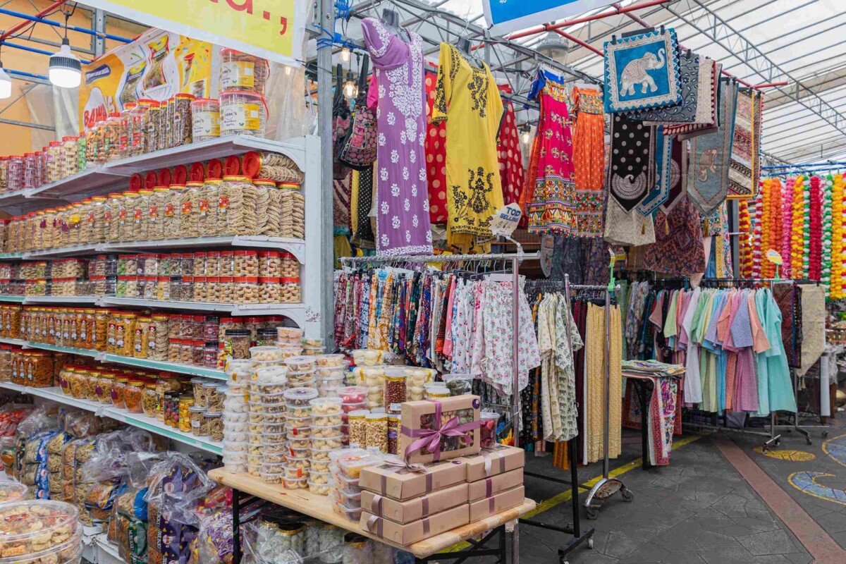 A Festival Village stall under a white metal roof, with shelves of sweets in clear jars, racks of patterned clothing and orange garlands hanging at the back